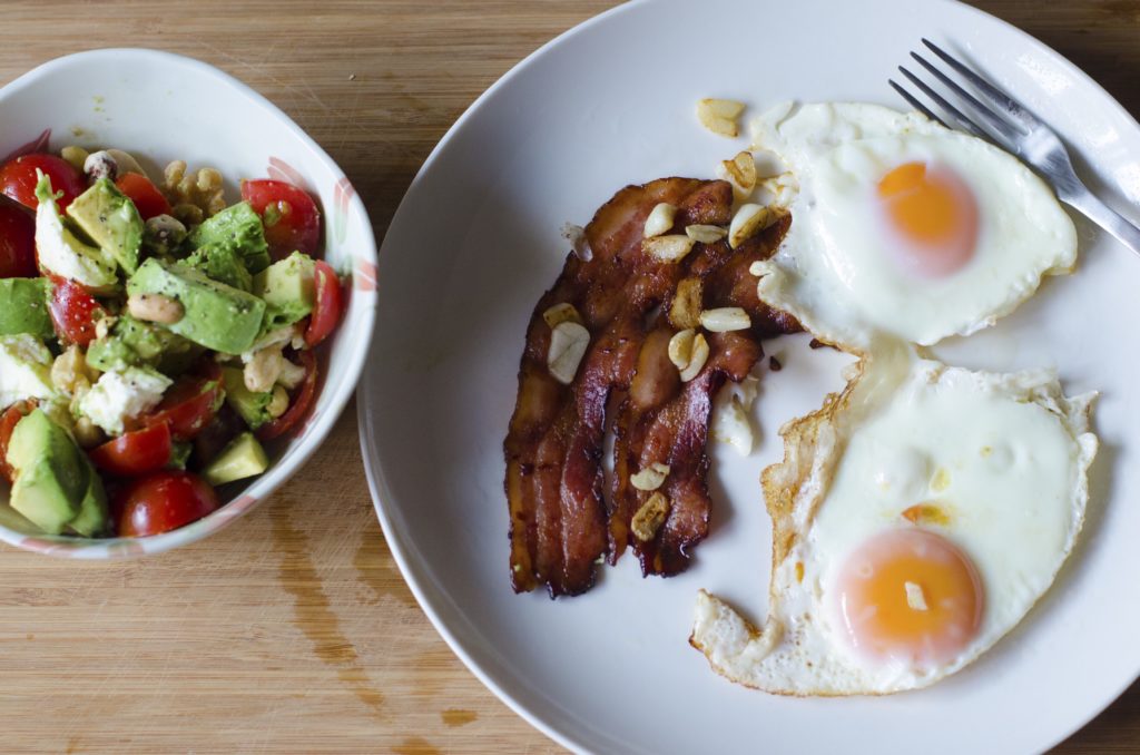 Two eggs sunny-side-up with two strips of bacon, next to a bowl of chopped avocado, tomato, and nuts